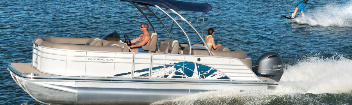 Man piloting a white 2020 Bennington® pontoon boat through the water as a woman looks out from …